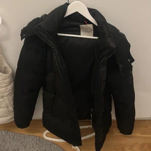Svart dunjacka från Moncler - Säljer en stilren svart dunjacka från Moncler. Jackan har en pufferdesign med dragkedja och hög krage för extra värme. Perfekt för kalla vinterdagar. Den har långa ärmar och en klassisk passform. En snygg och funktionell jacka för vintern.
