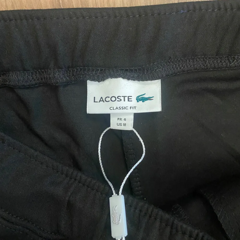Svarta mjukisbyxor från Lacoste med klassisk passform och dragsko i midjan. Byxorna har resår vid bensluten och en liten broderad Lacoste-logga på ena benet. Perfekta för en avslappnad stil.. Farkut & Housut.
