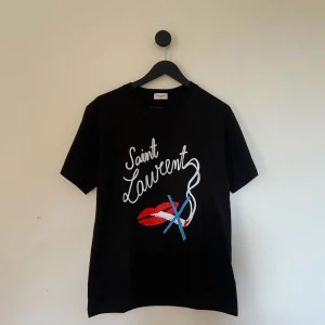 Saint Laurent T-shirt - Snygg svart t-shirt från Saint Laurent med ett coolt tryck av röda läppar och texten 'Saint Laurent' i vitt. Perfekt för sommarn!