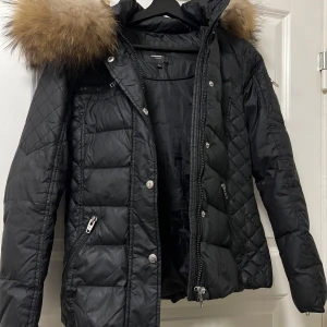 Svart dunjacka Rockandblue med päls - Snygg svart dunjacka från Rockandblue med fluffig päls runt huvan. Jackan har quiltade detaljer, dragkedjor och knappar framtill samt flera fickor. Perfekt för kalla dagar och ger en cool, urban vibe.