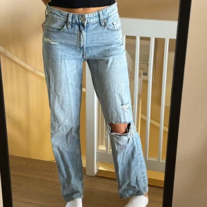 Ljusblå slitna straight jeans - Säljer ett par ljusblå jeans med raka ben och snygga slitningar, bland annat ett stort hål på ena knät. Klassisk femficksmodell med hög midja och avslappnad passform. Perfekta för en chill och trendig look.