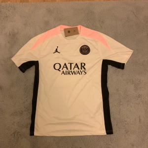 PSG x Jordan vit/svart fotbollströja - Snygg PSG fotbollströja från Jordan i vitt med svarta och ljusrosa detaljer. Kortärmad modell med svart Paris Saint-Germain-logga och Jordan-logga på bröstet. Tillverkad i lätt och ventilerande polyester, perfekt för träning eller match.
