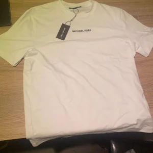 Vit t-shirt från Michael Kors XS - Vit t-shirt från Michael Kors i klassisk modell med rund hals och korta ärmar. Diskret svart logga broderad på bröstet. Tillverkad i mjuk bomull som känns skön mot huden. Perfekt för en clean och stilren look. Ny pris 899 kr säljs för 499