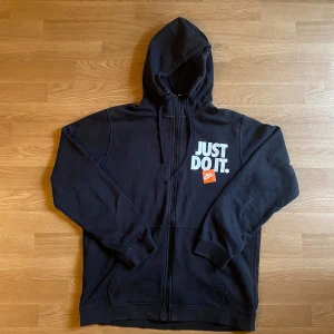 Svart Nike hoodie med tryck - Svart hoodie från Nike med dragkedja och huva. På bröstet finns det klassiska 'JUST DO IT.'-trycket i vitt och en orange Nike-logga. Tröjan har långa ärmar, kängurufickor och är gjord i mjuk bomullsmix. Perfekt för chill dagar eller när du vill ha en sportig look. Nypris 600kr