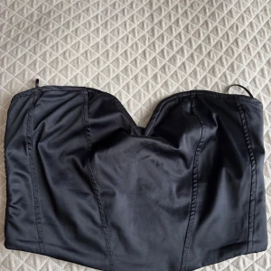 Svart satin korsett från Gina Tricot - Supersnygg svart korsett från Gina Tricot i glansigt satinmaterial. Toppen är axelbandslös med markerade sömmar och har en dragkedja bak för enkel påklädning. Perfekt för att skapa en trendig look med hög midja eller kjol.