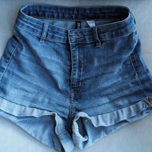 Jeansshorts från Divided, strl 34 - Snygga blå jeansshorts från Divided i klassisk denim med hög midja och uppvikta benslut. Shortsen har fickor både fram och bak samt bälteshällor. Perfekta för varma dagar och enkel att matcha med allt.