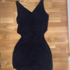 Svart omlottklänning med v-ringning - Snygg svart kortklänning med omlott och v-ringad hals. Ärmlös modell med markerad midja och smickrande passform. Perfekt för dig som gillar stilrena och enkla plagg med en modern twist. Vet ej storleken men runt S-M
