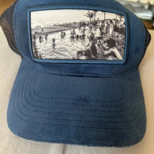 Blå truckerkeps Emilio Pendenza - Snygg blå truckerkeps från Emilio Pendenza med svart mesh baktill och justerbar snapback. Framsidan har en unik svartvit fotoprint av en strandscen. Kepsen har mjuk mockaliknande skärm och klassisk form.