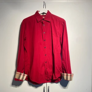Burberry skjorta - Snygg röd skjorta från Burberry med klassiska rutiga detaljer på insidan av kragen och ärmsluten. Skjortan har lång ärm, knappar framtill och broderad logga på bröstet. Perfekt för dig som vill sticka ut med en ikonisk look. Välanvänd men i fint skick. Storlek M. Nypris 5400kr. Sällsynt färg. Hör av er för fler bilder. 