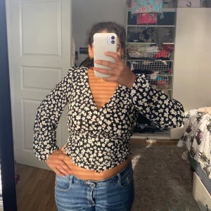 Svart blommig omlottopp med v-ringning - Säljer en svart omlottopp med vita blommor och v-ringning. Toppen har långa ärmar och smickrande passform med markerad midja. Perfekt för dig som gillar trendiga och feminina plagg med lite edge.