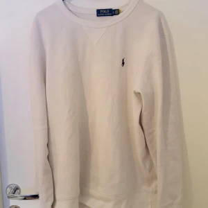 Vit sweatshirt från Polo Ralph Lauren - Klassisk vit sweatshirt från Polo Ralph Lauren med rund halsringning och broderad logga på bröstet. Tröjan har lång ärm och är tillverkad i mjuk bomull, perfekt för en clean och avslappnad stil. Liten fläck vid uriningen men går bort ifall man använder medel