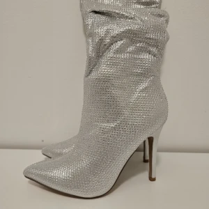 Glittriga silverstövletter med klack - Snygga stövletter i glittrigt silver med spetsig tå och hög klack. Skorna har en slouchy passform och är täckta av små glittrande stenar som ger en riktigt festlig look. Perfekta för dig som vill sticka ut och glänsa.