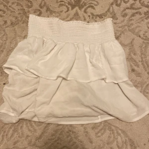 Vit volangkjol från lager 157 - Supersöt vit kortkjol från lager 157 med bred smock i midjan och dubbla volanger. Kjolen är lätt och luftig, perfekt för varma dagar. Enkel att matcha med crop tops eller en snygg hoodie för en chill look.