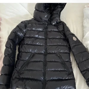 Moncler jacka bady  - Snygg svart pufferjacka från Moncler med glansig finish och huva. Jackan har dragkedja framtill, två snedställda fickor med dragkedja och klassisk Moncler-logga på ärmen. Perfekt för kalla dagar när du vill hålla stilen. Storlek 14 år men skulle absolut säga att den passar xs/s också!