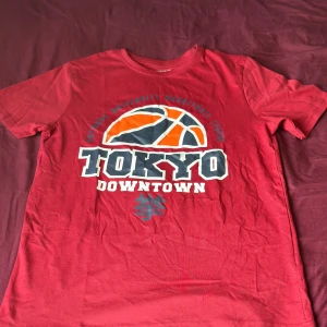 Röd Tokyo Downtown t-shirt - Cool röd t-shirt med stort tryck av en basketboll och texten 'Tokyo Downtown' på bröstet. T-shirten har klassisk passform och korta ärmar. Perfekt för dig som gillar sportig streetstyle och vill sticka ut med en färgstark look.