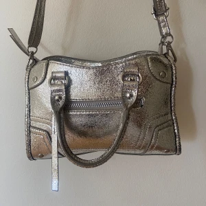 Silvrig handväska med axelrem - Knappt använd handväska i metallic silver med både handtag och justerbar axelrem. Väskan har en glansig, krackelerad yta och dragkedja upptill samt en ficka med dragkedja framtill. Perfekt storlek för det viktigaste och ger en riktigt trendig vibe 🌸