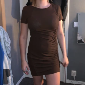 Brun kort klänning från H&M Divided - Säljer en tight brun kortklänning från H&M Divided i storlek S. Klänningen har korta ärmar, rund halsringning och snygg rynkdetalj med snörning på sidan. Materialet är stretchigt och följsamt, perfekt för dig som gillar en figurnära passform.