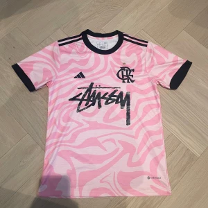 Rosa Adidas x Stüssy fotbollströja - Snygg rosa fotbollströja från Adidas i samarbete med Flamengo och Stüssy. Tröjan har korta ärmar, svart krage och muddar samt ett unikt abstrakt mönster i rosa nyanser. Tillverkad i lätt och ventilerande material, perfekt för träning eller streetwear. Storlek S och i riktigt fint skick