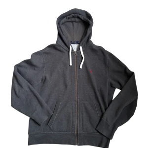 Polo Ralph Lauren - Mörkgrå hoodie från Polo Ralph Lauren med vit snörning i huvan och dragkedja framtill. Klassisk röd logga broderad på bröstet och två fickor framtill. Skön och avslappnad passform, perfekt för chill dagar.
