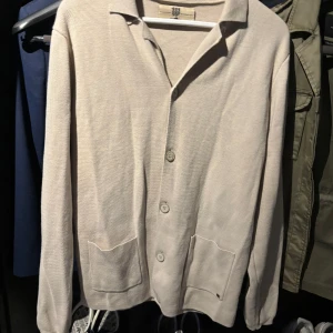 Beige cardigan från Bertoni i bomull - Stilren beige cardigan från Bertoni med klassisk krage, stora knappar och två praktiska fickor framtill. Cardiganen  är långärmad och tillverkad i mjuk bomull, perfekt för en clean och avslappnad look.