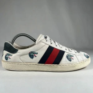 Gucci ace sneakers med vargmotiv - Gucci sneakers i vitt med ikoniska blå och röda ränder på sidorna. Skorna har tryckta varghuvuden över hela ovandelen och klassisk snörning. Snygg och unik design som sticker ut, perfekt för dig som vill ha något extra. Ingen box eller kvitto