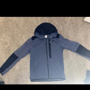 Blå Nike tech - Nike tech i blått med svarta detaljer och huva. Jackan har hel dragkedja framtill, bröstficka med dragkedja och sportig look. Perfekt för dig som gillar streetwear och vill ha något stilrent men ändå funktionellt.