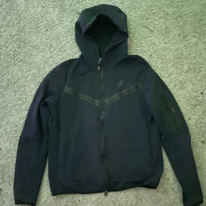 Mörkblå Nike tech kofta med huva - Mörkblå Nike tech kofta med hel dragkedja och huva. Koftan har en diskret Nike-logga på bröstet och en extra ficka med dragkedja på ena ärmen. Perfekt för dig som gillar sportig och clean stil.