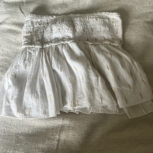 Vit kortkjol med guldiga detaljer - Supersöt vit kortkjol med smockad midja och volangig, luftig kjol. Luftigt tyg med små guldiga inslag som ger en extra touch. Perfekt för varma dagar och somriga vibes. Har inbyggda shorts och sitter bekvämt i midjan. Köpt utomlands 