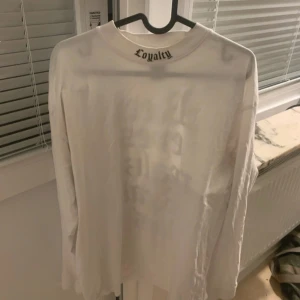 Vit långärmad tröja Black Squad S - Vit långärmad tröja från Black Squad med trycket 'Loyalty' i nacken och stort svart texttryck på ryggen. Relaxed fit och crewneck. Perfekt för dig som gillar streetwear och statement-plagg.