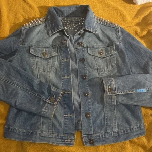Jeansjacka med nitar från DS.JEANS - Snygg blå jeansjacka från DS.JEANS med coola silvriga nitar över axlar och övre rygg. Klassisk modell med knappar framtill, bröstfickor och långa ärmar. Perfekt för dig som vill sticka ut med en edgy vibe.