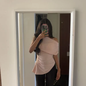 Babyrosa topp - Säljer en ljusrosa asymmetrisk topp med oneshoulder och rynkade detaljer på sidan. Toppen har en cool drapering och är figurnära med en snygg sned nederkant. Perfekt för dig som gillar unika snitt och mjuka material.