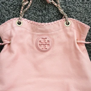 Väska från Tory Burch - Supersöt rosa axelväska från Tory Burch i mjukt skinn med kedjedetaljer i axelbandet och snyggt flätad kant upptill. Väskan har det klassiska Tory Burch-logot på framsidan och dragkedjefack inuti. Perfekt storlek för allt du behöver ha med dig.