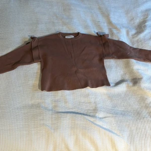 Brun tröja med rosetter från Zara - Säljer en brun långärmad tröja från Zara med söta rosetter på axlarna. Tröjan har en enkel, rak passform och är gjord i ett mjukt material som känns skönt mot huden. Perfekt för dig som gillar stilrena detaljer och en chill look.