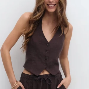 Brun linneväst med knappar - Snygg brun väst i linne med v-ringning och fyra knappar framtill. Modellen är croppad och har en klassisk, figurnära passform som ger en stilren look. Perfekt att styla över en t-shirt eller direkt mot huden för en trendig vibe. Aldrig använd, prislappar kvar. Nypris 399kr