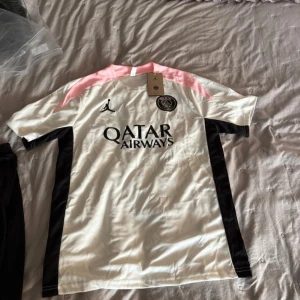 PSG x Jordan vit fotbollströja M - Säljer en PSG x Jordan fotbollströja i vitt med svarta detaljer och rosa axelparti. Tröjan har korta ärmar, broderad PSG-logga och Jordan-logga på bröstet samt Qatar Airways-tryck. Tillverkad i lätt och ventilerande material, perfekt för match eller träning.