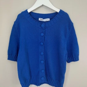 Blå stickad kofta från Zara - Superfin blå stickad kofta från Zara med korta ärmar och stora klädda knappar framtill. Koftan har ribbade muddar vid ärmslut och nederkant, perfekt för lager-på-lager och en poppig look.