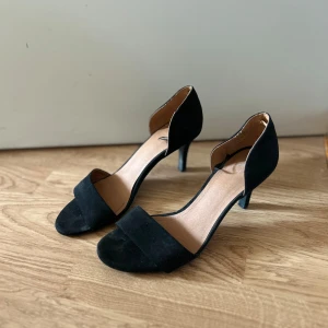 Svarta pumps med klack och öppen tå - Stilrena svarta pumps i mocka med öppen tå och låg klack. Snygg minimalistisk design med bred rem över tårna och stängd häl. Perfekta för att lyfta din outfit med en elegant touch.