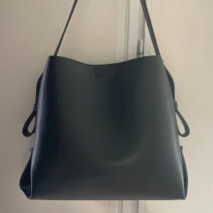 Svart axelväska i skinn - Stilren svart axelväska i slätt skinn med minimalistisk design. Väskan har justerbara bältesdetaljer på sidorna och ett stort huvudfack med dragkedja. Perfekt för dig som vill ha en enkel men snygg väska med mycket plats. Inga tecken på användning, har glömt vart jag köpte den då lappen är borta men nypris ca 400