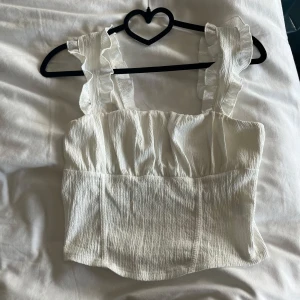 Vit linnetopp med volangband - Supersöt vit linnetopp med breda axelband och volangdetaljer. Toppen har en lätt rynkad struktur och är sydd i ett luftigt tyg som passar perfekt till varma dagar. Snygg fyrkantig halsringning och figurnära passform.