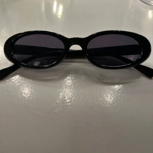 Svarta ovala solglasögon från Bershka - Snygga svarta solglasögon från Bershka med ovala bågar och mörka glas. Ramen är i blank plast och ger en cool retrovibe. Perfekta för att lyfta din stil med en touch av 90-talskänsla.