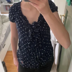 Mörkblå prickig blus från Hollister XS - Supersöt mörkblå blus från Hollister med vita prickar, volangdetaljer längs knäppningen och lätt puffade korta ärmar. Blusen har knappar framtill och en liten broderad logga vid höften. Perfekt för dig som gillar en feminin och lekfull stil.