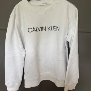 Vit sweatshirt från Calvin Klein - ungdom - Vit sweatshirt från Calvin Klein med svart logotyptryck framtill. Klassisk rund halsringning, ribbade muddar och lång ärm. Perfekt för en clean och stilren look.