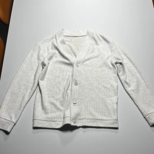 Vit cardigan  - Säljer en stilren vit cardigan med ribbad struktur och fyra knappar framtill. Koftan har långärmad passform och är perfekt att slänga på sig över en t-shirt. Enkel och clean look som funkar till det mesta.