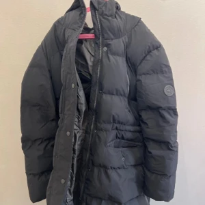 Svart pufferjacka från Everest - Svart pufferjacka från Everest med huva och dragkedja samt knappar framtill. Jackan har två fickor med lock och en diskret logga på ärmen. Perfekt för kalla dagar och riktigt skön att bära. Klassisk och enkel design som funkar till det mesta.