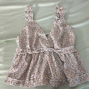 Blommig linnetopp från SHEIN - Supersöt blommig linnetopp från SHEIN i vitt med små rosa, gröna och blå detaljer. Toppen har volangkant, breda axelband och smockad rygg för extra komfort. Perfekt för varma dagar och ger en romantisk vibe till din outfit.