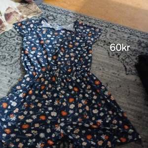 Blommig kortklänning i marinblått - Supersöt kortklänning med marinblå botten och blommigt mönster i vitt, orange och beige. Klänningen har korta ärmar och resår i midjan för en skön passform. Perfekt för varma dagar och somriga vibes!