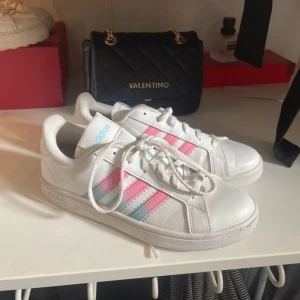 Adidas sneakers med rosa och blå ränder - Snygga vita Adidas sneakers med klassiska tre ränder i rosa och ljusblått på sidorna. Skorna har snörning, rund tå och platt sula. Skorna är näst intill aldrig använda, pris kan diskuteras 