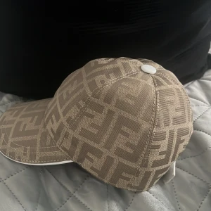 Beige Fendi keps med FF-mönster - Snygg beige keps från Fendi med ikoniskt FF-logomönster över hela kepsen. Justerbar rem bak i vitt läder med Fendi-logga. Tillverkad i bomull och polyester, perfekt för dig som vill sticka ut med en lyxig accessoar.