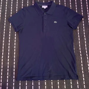 Mörkblå Lacoste pikétröja slim fit - Säljer en mörkblå pikétröja från Lacoste i slim fit-modell. Klassisk krage, tre knappar framtill och den ikoniska gröna krokodilloggan på bröstet. Tillverkad i mjuk bomull, perfekt för en clean och sportig look.