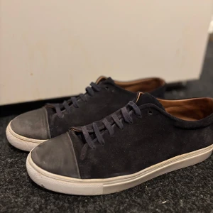 Mörkblå mocka skor  - Stilrena svarta sneakers i mocka med rund tå och vit platt sula. Skorna har svarta snören och en diskret grå tådel som ger en cool kontrast. Insidan är fodrad i brunt för en snygg touch. Perfekta för dig som gillar minimalistisk och clean stil.